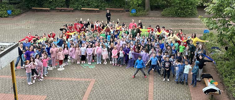Kinder der Grundschule am Kreuzweg formen bunt verkleidet einen Regenbogen auf dem Schulhof